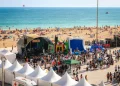 Un vivace scenario dell' Albufeira Sea Fest con persone radunate attorno a un palco, attrazioni gonfiabili e tende bianche. La folla si gode l'evento vicino a una spiaggia sabbiosa con bagnanti e bagnanti in riva al mare blu scintillante.