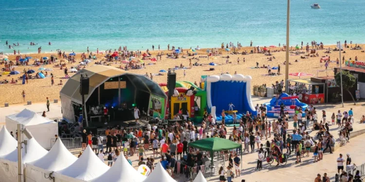 Un vivace scenario dell' Albufeira Sea Fest con persone radunate attorno a un palco, attrazioni gonfiabili e tende bianche. La folla si gode l'evento vicino a una spiaggia sabbiosa con bagnanti e bagnanti in riva al mare blu scintillante.