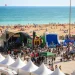 Un vivace scenario dell' Albufeira Sea Fest con persone radunate attorno a un palco, attrazioni gonfiabili e tende bianche. La folla si gode l'evento vicino a una spiaggia sabbiosa con bagnanti e bagnanti in riva al mare blu scintillante.