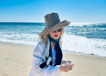 Madonna avvistata con un grande cappello da sole, occhiali da sole e una giacca bianca è in piedi su una spiaggia sabbiosa di Comporta a Lisbona con l'oceano e un cielo azzurro brillante sullo sfondo.
