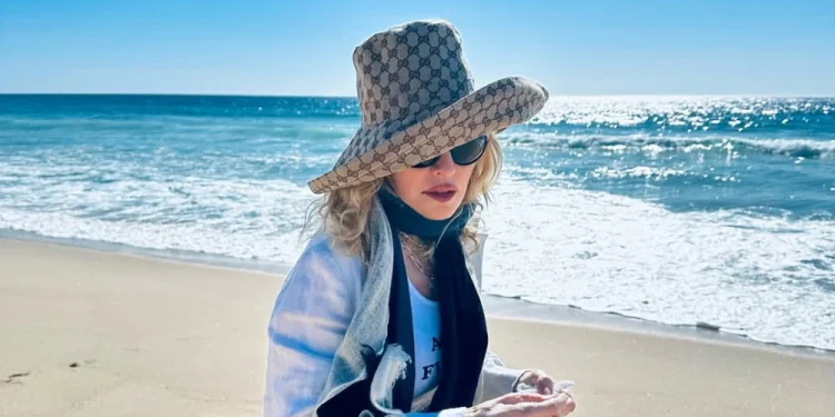 Madonna avvistata con un grande cappello da sole, occhiali da sole e una giacca bianca è in piedi su una spiaggia sabbiosa di Comporta a Lisbona con l'oceano e un cielo azzurro brillante sullo sfondo.