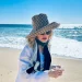 Madonna avvistata con un grande cappello da sole, occhiali da sole e una giacca bianca è in piedi su una spiaggia sabbiosa di Comporta a Lisbona con l'oceano e un cielo azzurro brillante sullo sfondo.