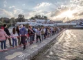 Un folto gruppo di persone passeggia lungo un ponte pedonale galleggiante durante il Festival do Contrabando di Alcoutim che attraversa il Guadiana. Il sole tramonta sullo sfondo, gettando raggi attraverso le nuvole, edifici e alberi costeggiano la riva del fiume sotto un cielo parzialmente nuvoloso.