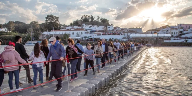 Un folto gruppo di persone passeggia lungo un ponte pedonale galleggiante durante il Festival do Contrabando di Alcoutim che attraversa il Guadiana. Il sole tramonta sullo sfondo, gettando raggi attraverso le nuvole, edifici e alberi costeggiano la riva del fiume sotto un cielo parzialmente nuvoloso.
