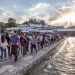 Un folto gruppo di persone passeggia lungo un ponte pedonale galleggiante durante il Festival do Contrabando di Alcoutim che attraversa il Guadiana. Il sole tramonta sullo sfondo, gettando raggi attraverso le nuvole, edifici e alberi costeggiano la riva del fiume sotto un cielo parzialmente nuvoloso.