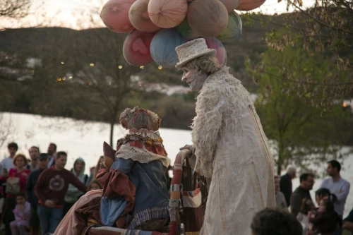 Due artisti in costume con abiti in stile vintage onorano il palco del Festival Contrabando Alcoutim. Uno con cappello e abito bianco è in piedi mentre l'altro con un vestito colorato è seduto. Sono vicino a una folla, vicino all'acqua, circondati da alberi, palloncini e la collina ondulata sullo sfondo.