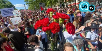 Un frame del video del 25 aprile a Lisbona, dove grande folla si raduna all'aperto durante il giorno, molti con cartelli e garofani rossi. Una mano con un mazzo di garofani rossi spicca in primo piano, con alberi ed edifici sullo sfondo.