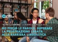 Un gruppo di ragazzi, superando le difficoltà della barriera linguistica, siedono in un ambiente informale al chiuso, con persone che parlano mentre altri ascoltano attentamente. Scaffali pieni di libri sono sullo sfondo, a sottolineare la loro ricerca di superare la frustrazione e condividere i loro sentimenti.