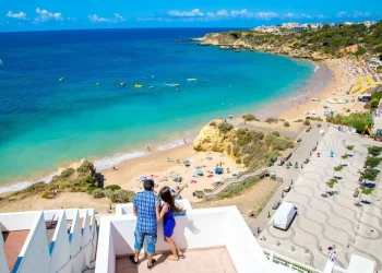 Una coppia è in piedi su un balcone che si affaccia su una pittoresca spiaggia del Portogallo, con acqua turchese, sabbia dorata, bagnanti e un lungomare, forse chiedendosi quale sia il costo della camera albergo per la loro fuga perfetta sotto il limpido cielo azzurro.