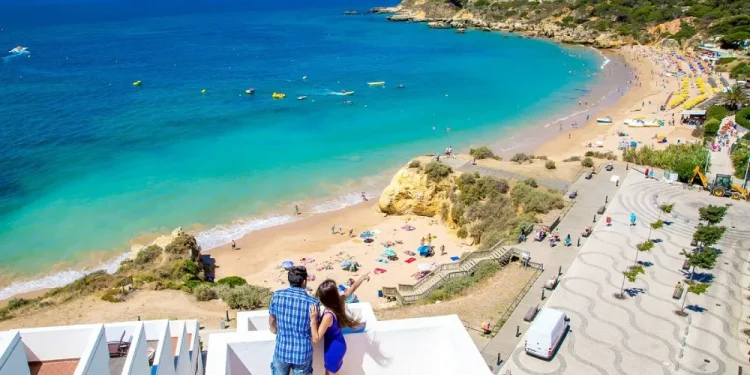 Una coppia è in piedi su un balcone che si affaccia su una pittoresca spiaggia del Portogallo, con acqua turchese, sabbia dorata, bagnanti e un lungomare, forse chiedendosi quale sia il costo della camera albergo per la loro fuga perfetta sotto il limpido cielo azzurro.