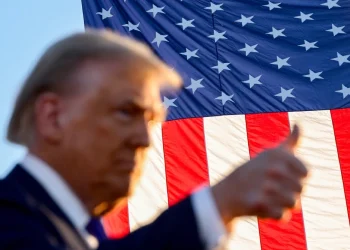 Dopo gli effetti dei dazi in Portogallo, Donald Trump fa il gesto del pollice in su in primo piano, leggermente fuori fuoco. Sullo sfondo, una grande bandiera americana sventola in modo evidente contro un cielo azzurro terso.