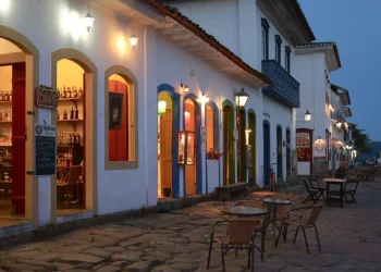 Scena di strada al tramonto senza lavoratori e senza rischio in Portogallo con una fila di negozi illuminati in edifici in stile coloniale. La strada acciottolata è fiancheggiata da tavoli e sedie vuoti all'aperto. Il cielo è di un blu sempre più intenso e luci calde illuminano gli ingressi dei negozi.
