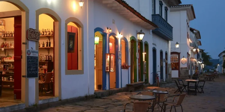 Scena di strada al tramonto senza lavoratori e senza rischio in Portogallo con una fila di negozi illuminati in edifici in stile coloniale. La strada acciottolata è fiancheggiata da tavoli e sedie vuoti all'aperto. Il cielo è di un blu sempre più intenso e luci calde illuminano gli ingressi dei negozi.
