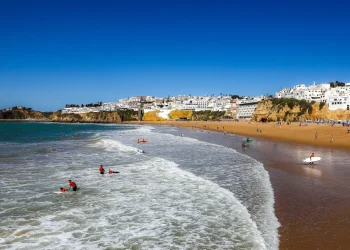 Persone fanno surf e nuota tra le onde su una spiaggia sabbiosa con una città costiera con edifici bianchi e scogliere sullo sfondo, godendo di un clima perfetto, come mostra Meteo Portogallo, sotto un cielo azzurro e limpido.