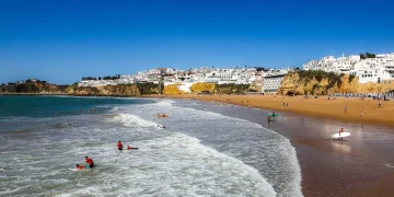 Persone fanno surf e nuota tra le onde su una spiaggia sabbiosa con una città costiera con edifici bianchi e scogliere sullo sfondo, godendo di un clima perfetto, come mostra Meteo Portogallo, sotto un cielo azzurro e limpido.