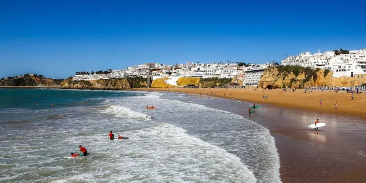 Persone fanno surf e nuota tra le onde su una spiaggia sabbiosa con una città costiera con edifici bianchi e scogliere sullo sfondo, godendo di un clima perfetto, come mostra Meteo Portogallo, sotto un cielo azzurro e limpido.