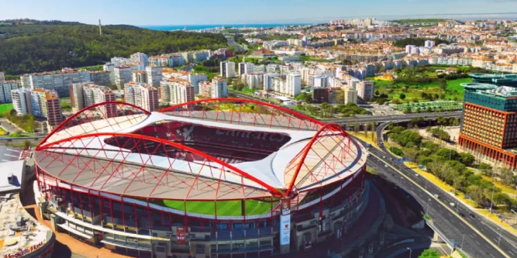Vista aerea dell'iconico stadio da Luz Benfica in trattativa con Allianz, un grande stadio di calcio con una caratteristica struttura in acciaio rosso, circondato da edifici urbani, strade e vegetazione sotto un cielo azzurro e terso.