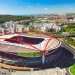 Vista aerea dell'iconico stadio da Luz Benfica in trattativa con Allianz, un grande stadio di calcio con una caratteristica struttura in acciaio rosso, circondato da edifici urbani, strade e vegetazione sotto un cielo azzurro e terso.