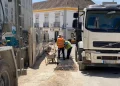 In un cantiere edile portoghese, due operai, con reddito annuo garantito e con giubbotti di sicurezza arancioni e gialli sono in piedi tra due grossi camion . Lì vicino, una carriola aspetta di essere utilizzata. Sullo sfondo, un edificio con balconi si crogiola sotto il cielo soleggiato.