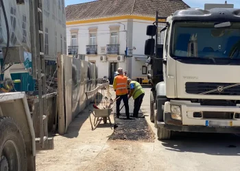 In un cantiere edile portoghese, due operai, con reddito annuo garantito e con giubbotti di sicurezza arancioni e gialli sono in piedi tra due grossi camion . Lì vicino, una carriola aspetta di essere utilizzata. Sullo sfondo, un edificio con balconi si crogiola sotto il cielo soleggiato.