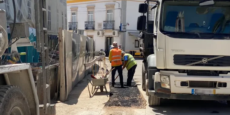 In un cantiere edile portoghese, due operai, con reddito annuo garantito e con giubbotti di sicurezza arancioni e gialli sono in piedi tra due grossi camion . Lì vicino, una carriola aspetta di essere utilizzata. Sullo sfondo, un edificio con balconi si crogiola sotto il cielo soleggiato.