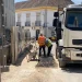 In un cantiere edile portoghese, due operai, con reddito annuo garantito e con giubbotti di sicurezza arancioni e gialli sono in piedi tra due grossi camion . Lì vicino, una carriola aspetta di essere utilizzata. Sullo sfondo, un edificio con balconi si crogiola sotto il cielo soleggiato.