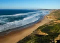 Vista panoramica di una delle 9 spiagge portoghesi ricche di iodio: quella di Vale Figueiras dove l'oceano con le onde si infrangono. Questa praia presenta un litorale costeggiato da vegetazione verde e scogliere frastagliate sotto un cielo azzurro e limpido, che si estende a perdita d'occhio.