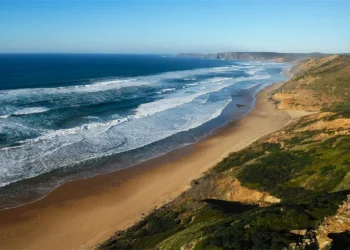 Vista panoramica di una delle 9 spiagge portoghesi ricche di iodio: quella di Vale Figueiras dove l'oceano con le onde si infrangono. Questa praia presenta un litorale costeggiato da vegetazione verde e scogliere frastagliate sotto un cielo azzurro e limpido, che si estende a perdita d'occhio.