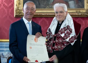 Sergio Mattarella e Marcelo Rebelo de Sousa sono in piedi l'uno accanto all'altro in occasione della consegna del Honoris Causa a Coimbra. Uno, avvolto in una tunica decorata con dettagli rossi e argento, presenta l'attestato. Si trovano in una stanza elegantemente decorata con carta da parati rossa e un dipinto incorniciato all'Università di Coimbra.