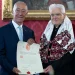 Sergio Mattarella e Marcelo Rebelo de Sousa sono in piedi l'uno accanto all'altro in occasione della consegna del Honoris Causa a Coimbra. Uno, avvolto in una tunica decorata con dettagli rossi e argento, presenta l'attestato. Si trovano in una stanza elegantemente decorata con carta da parati rossa e un dipinto incorniciato all'Università di Coimbra.
