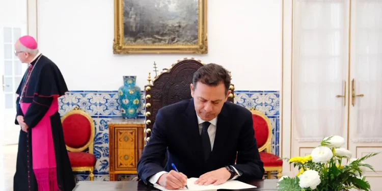 Luís Montenegro firma un documento, probabilmente in relazione alle politiche di espulsione degli immigrati irregolari in Portogallo, seduto a un tavolo di legno decorato con fiori bianchi, mentre un sacerdote osserva sullo sfondo di una sala formale.