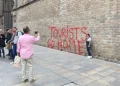 Un gruppo di persone scatta foto vicino a un grande muro in Spagna con graffiti rossi che recitano "TURISTI TORNATE A CASA", a simboleggiare le proteste contro il turismo di massa. Una donna posa accanto al messaggio, mentre altre sono in piedi accanto a un muro in pietra all'aperto.