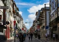Una vivace via pedonale a Porto fiancheggiata da edifici colorati e balconi che si colloca in alto nella classifica affitti delle città più care del Portogallo, mentre le persone passeggiano lungo l'ampio sentiero piastrellato sotto un cielo azzurro brillante con nuvole sparse.