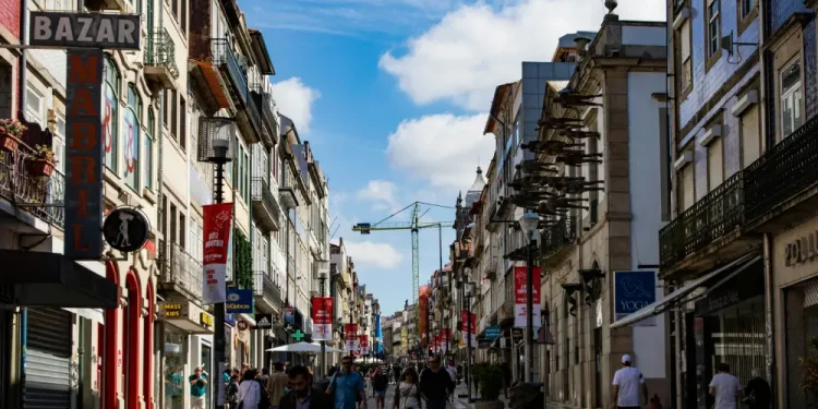 Una vivace via pedonale a Porto fiancheggiata da edifici colorati e balconi che si colloca in alto nella classifica affitti delle città più care del Portogallo, mentre le persone passeggiano lungo l'ampio sentiero piastrellato sotto un cielo azzurro brillante con nuvole sparse.