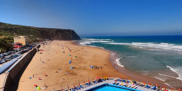 Praia Grande a Sintra in Portogallo si estende lungo la costa, accanto a una grande piscina all'aperto circondata da lettini colorati, perimata con Bandiera Blu nel 2025. Onde che si infrangono e persone che si rilassano sotto un cielo azzurro e limpido.