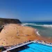 Praia Grande a Sintra in Portogallo si estende lungo la costa, accanto a una grande piscina all'aperto circondata da lettini colorati, perimata con Bandiera Blu nel 2025. Onde che si infrangono e persone che si rilassano sotto un cielo azzurro e limpido.