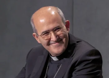 Il papabili cardinale portoghese Tolentino de Mendonça, con gli occhiali, che indossa un abito clericale nero e un colletto clericale bianco, sorride mentre guarda leggermente di lato su uno sfondo grigio semplice.