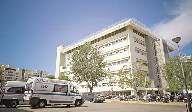 L'ospedale di Faro in Algarve dove si cercano nuovi medici, con alcune ambulanze parcheggiate vicino a tre alberi. Il cielo è azzurro con alcune case sullo sfondo.