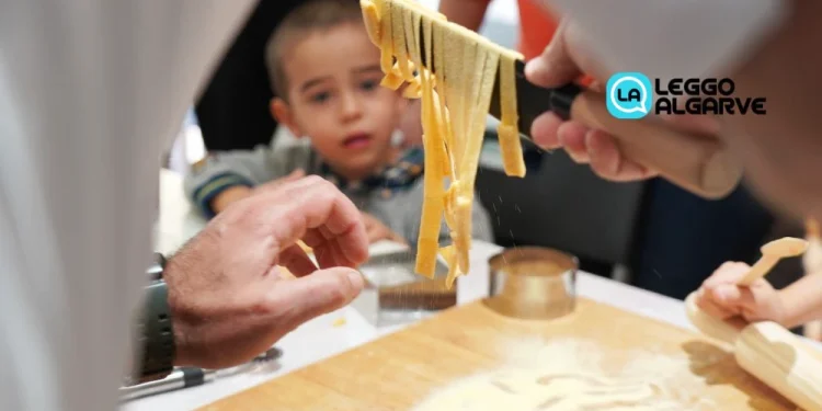 Un primo piano di mani che preparano la pasta fresca su una superficie di legno infarinata durante un evento di pasta fresca in Algarve, con un bambino che osserva attentamente sullo sfondo. Il logo di Leggo Algarve è visibile nell'angolo in alto a destra.