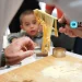Un primo piano di mani che preparano la pasta fresca su una superficie di legno infarinata durante un evento di pasta fresca in Algarve, con un bambino che osserva attentamente sullo sfondo. Il logo di Leggo Algarve è visibile nell'angolo in alto a destra.