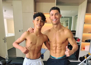 Crisitiano Ronaldo con il figlio a torso nudo sono in piedi, vicini in una palestra, sorridenti verso la telecamera. Entrambi mostrano addominali scolpiti e fanno il gesto del pollice in su. Sullo sfondo si vedono attrezzi da palestra e scaffali.