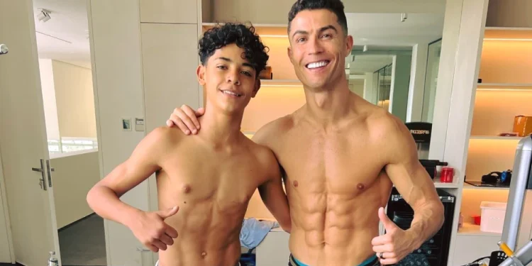 Crisitiano Ronaldo con il figlio a torso nudo sono in piedi, vicini in una palestra, sorridenti verso la telecamera. Entrambi mostrano addominali scolpiti e fanno il gesto del pollice in su. Sullo sfondo si vedono attrezzi da palestra e scaffali.
