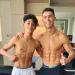 Crisitiano Ronaldo con il figlio a torso nudo sono in piedi, vicini in una palestra, sorridenti verso la telecamera. Entrambi mostrano addominali scolpiti e fanno il gesto del pollice in su. Sullo sfondo si vedono attrezzi da palestra e scaffali.