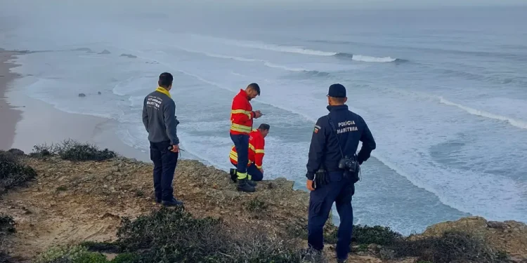 Quattro soccorritori, tra cui vigili del fuoco e agenti di polizia marittima, sono in piedi su una scogliera rocciosa che domina l'oceano, durante la ricerca dei due fratelli scomparsi in mare sulla spiaggia sottostante in una giornata nuvolosa.