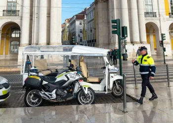 Un tuk-tuks parcheggiato a Lisbona e un poliziotto che si accinge ad emettere la multa. Si vede anche una moto e dei palazzi antichi sullo sfondo.