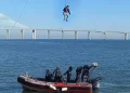 Una persona viene calata con un cavo dalla funivia di Lisbona, sopra una barca di salvataggio in acqua, con tre membri dell'equipaggio a bordo. Sullo sfondo, un lungo ponte Vasco da Gama che si estende sotto un cielo terso e azzurro.