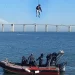 Una persona viene calata con un cavo dalla funivia di Lisbona, sopra una barca di salvataggio in acqua, con tre membri dell'equipaggio a bordo. Sullo sfondo, un lungo ponte Vasco da Gama che si estende sotto un cielo terso e azzurro.