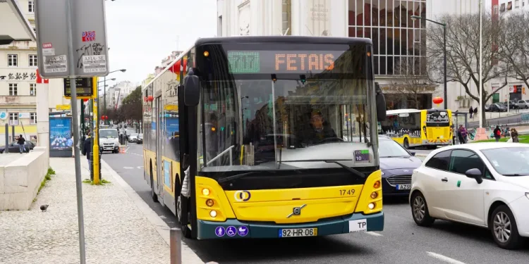 Un autobus dei trasporti urbano giallo sulla linea 717 per Fetais percorre una strada di Lisbona, in Portogallo, tra auto e pedoni, attualmente lo si deve pagare, ma in futuro potrebbe essere gratuiti. Sono visibili edifici, un teatro e cartelli stradali.
