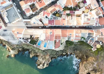 Vista aerea di case sul mare con tetti in terracotta lungo una costa rocciosa in Algarve, dove le onde si infrangono sotto e una strada con macchine parcheggiate corre parallela alla riva, rivelando il fascino che spesso nasconde rischi come una truffa immobiliare.