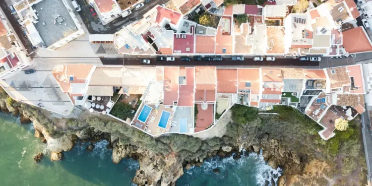 Vista aerea di case sul mare con tetti in terracotta lungo una costa rocciosa in Algarve, dove le onde si infrangono sotto e una strada con macchine parcheggiate corre parallela alla riva, rivelando il fascino che spesso nasconde rischi come una truffa immobiliare.